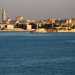 Sulla nostra sinistra appare lo skyline del centro storico di Bari, che sembra quasi galleggiare sul mare calmo e blu. In primo piano scorgiamo lottocentesco faro borbonico, mentre alle sue spalle si mostrano i massicci torrioni del Castello e la Cattedrale, con il suo alto campanile